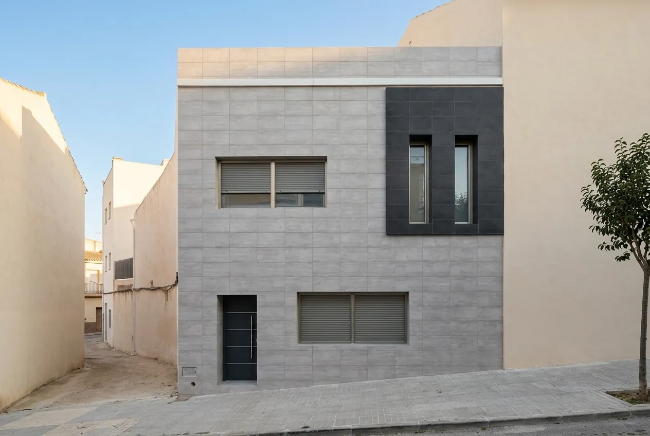 Vivienda entre medianeras de dos plantas, con fachada cerámica gris y un paño negro con dos ventanas verticales